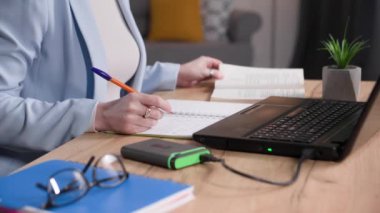 Genç bir kadın not defterine not alıyor. Evdeki masada dizüstü bilgisayarla çalışıyor.