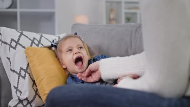 子供の健康少女は枕の上に寝そべって母親に喉を開けてソファの上に寝そべってる — ストック動画