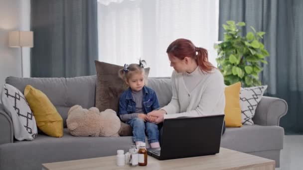 かわいい病気の娘を持つ若い女性は自宅のソファに座っている間 薬の選択についてのノートパソコンのコンサルティングでビデオリンクを介して医師と話しています — ストック動画