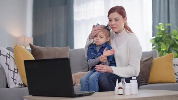 家庭でソファに座っている間にラップトップのビデオリンクで小児科医に相談している小さな女性の子供と一緒にオンラインの医者 若い母親は風邪やウイルスの治療を選択します — ストック動画