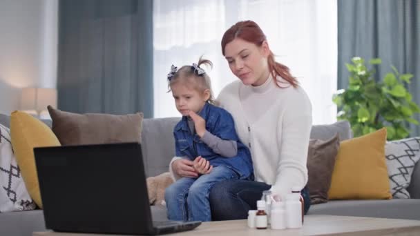 現代医学 女性の赤ん坊を持つ女性の腕の中で医師とオンラインでビデオ通話で話している自宅のソファに座ってラップトップでビデオ通話を使用して — ストック動画