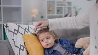 Virüs ya da enfeksiyon yüzünden boğazı ağrıyan sevimli bir kadın çocuk kanepede yatıyor, şefkatli bir anne, sağlığıyla ilgilenen kızının başını okşuyor.