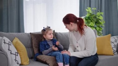 Çocuk sağlığı için endişelenen kadın, küçük bir kızla sağlık durumu hakkında konuşuyor, çocuğun kafasını avuç içi ile okşuyor, kanepede otururken sıcaklığı kontrol ediyor.