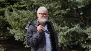 Beyaz saçlı, gözlüklü, elinde balta olan yaşlı bir adamın portresi. Bir ladin arka planında gülümseyen ve kameraya bakan.