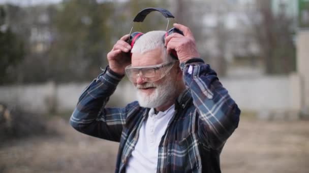 Retrato Anciano Gafas Construcción Con Auriculares Sonrientes Fondo Árboles — Vídeo de stock