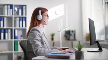 Gözlüklü hoş bir kadın çağrı merkezi operatörü olarak çalışıyor ve ofiste bilgisayar başında otururken kulaklık kullanarak müşteriyle konuşuyor.