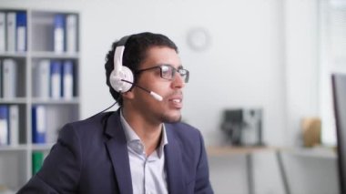 Teknik destek, afro amerikan erkek ofis çalışanı ofiste bilgisayar başında otururken kulaklık kullanarak müşteriyle konuşuyor.