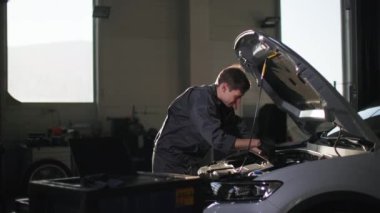Otomobil servisi, ingiliz anahtarı tamir eden üniformalı genç adam güneş ışığı peronunda açık bir kaputun altında motoru tamir ediyor.
