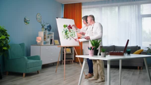 引退したエンターテイメント 才能ある老人と女性は 自宅でブラシと塗料でキャンバスに現代的な絵を描きます — ストック動画