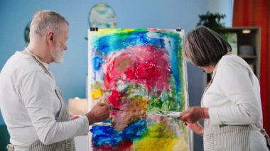 Emekli olan yaşlı erkek ve kadın emekliler, rahat bir odada sehpa kullanarak tuvale boya ve fırçayla resim çiziyorlar.