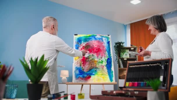 新しいスキル 才能のあるシニア女性と男性は自宅でイーゼルとブラシやキャンバスで塗料を使用して退職でペイントすることを学ぶ — ストック動画