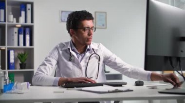 Gözlüklü zenci bir erkek doktorun portresi. Fonendoskopla röntgenleri incelerken, muayenehanede bilgisayarın başında oturuyor.