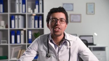 Online tıp, fonoendoskoplu genç profesyonel doktor ofiste otururken bilgisayardaki video bağlantısı ile hastayla konuşuyor, kameraya bakıyor.