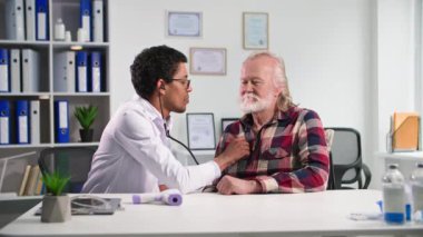 Muayenehanedeki hastayı muayene eden genç siyahi bir doktor, ofisinde fonendoskopla yaşlı bir adamın nefes alışını dinliyor.