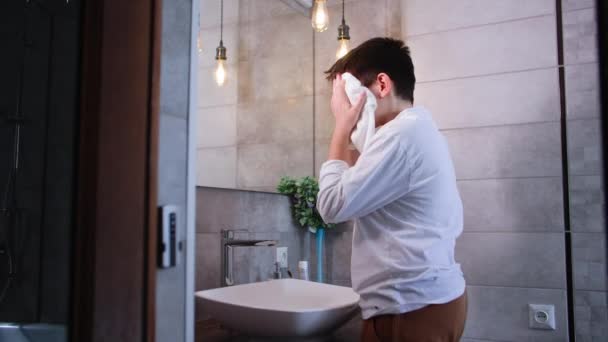 かわいい少年は冷たい水で顔を洗い 浴室の洗面台の近くに立っている間タオルで顔を拭きます — ストック動画