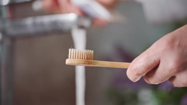 朝の儀式 洗面台近くの水でタップの歯ブラシの背景に歯磨き粉を塗る少年 クローズアップ — ストック動画
