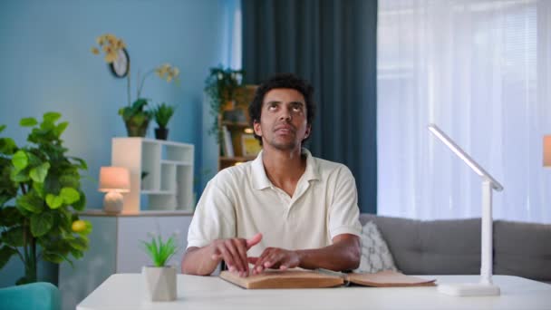 目が不自由な若い黒人男性は 部屋のテーブルに座って文字でページの上に指を走らせているブレイユの本を読んで — ストック動画