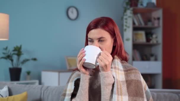 따뜻한 플레이드 미소와 카메라를 소파에 뜨거운 마시는 매력적인 여자의 초상화 — 비디오