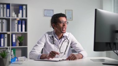 Profesyonel siyah erkek doktor hastalarla bilgisayardaki video bağlantısıyla iletişim kuruyor. Tıp ofisinde otururken dahili bir web kamerası kullanıyor.