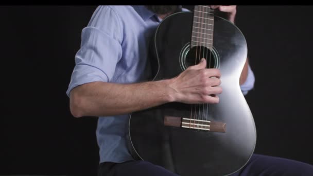 ギターを演奏する男性ミュージシャンは 彼の手で楽器を保持し 暗い背景に座っている間 メロディーを演奏する弦をプルック クローズアップ — ストック動画