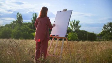 Fırça ve akrilik boya kullanan yaratıcı kadın ressam açık havada bir resim çiziyor.