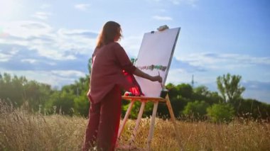 Elinde fırça ve palet olan yaratıcı kadın, mavi güneş ışınları gökyüzüne karşı, doğa sehpasını kullanarak tuval üzerine bir resim çiziyor.