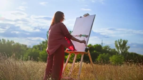 一个富有创造力的女人 手里拿着画笔和调色板 用画架在画布上 在蓝天的阳光下 画一幅画 — 图库视频影像
