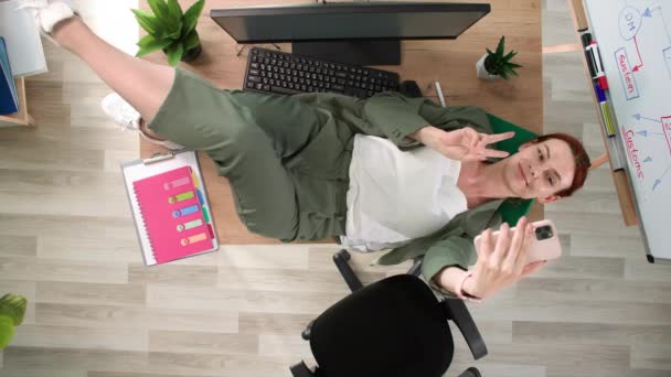 マネージャーの勤務日 若い面白い女性はコーヒー休憩中にテーブルの上に横たわり オフィスの電話でセルフィーを取ります トップビュー — ストック動画
