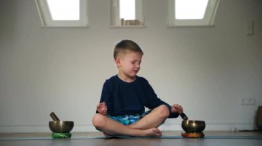 Erkek çocuk meditasyon yaparken müzik kaseleriyle oynuyor ve odada karikatür üzerinde otururken yoga yapıyor.