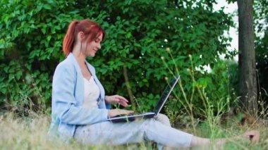 Serbest çalışan genç bir ofis çalışanı. Parktaki ağaçların çimlerinde otururken dizüstü bilgisayarla çalışıyor.