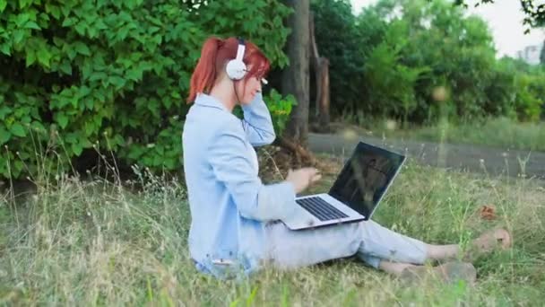 在室外工作时 年轻的女性呼叫中心员工坐在公园的草地上 使用耳机 并通过笔记本电脑上的视频链接进行交流 — 图库视频影像