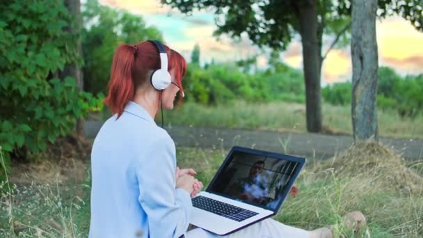여자는 나무의 배경에서 클리어링에 앉아있는 노트북에 헤드폰과 비디오 통신을 보스와 — 비디오