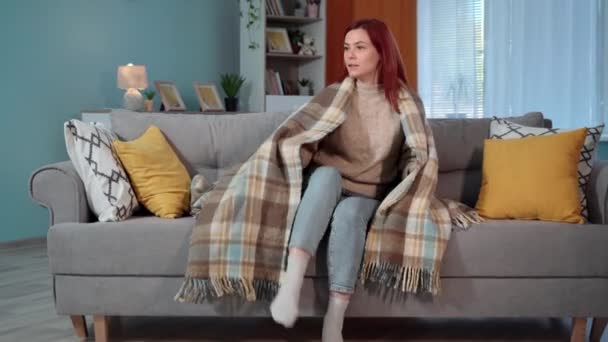 暖かい毛布の主婦は家の暖房の欠如のために家で冷たいのでソファーに座ります — ストック動画