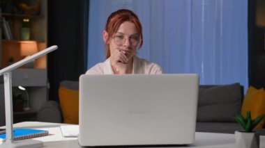 Ev ofisi, gözlüklü düşünceli genç bir kadın masada otururken bilgisayar başında çalışıyor.