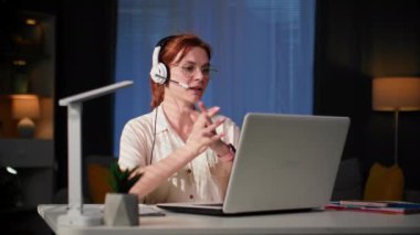 Ev ofisi, gözlüklü genç bir kadın patronuyla kulaklık ve video iletişimiyle online iletişim kuruyor ve masasında otururken bir not defterine not alıyor.