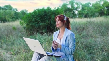 Dışarıda çalışan, kulaklıklı genç bir kadın yeşil çalıların arka planında otururken dizüstü bilgisayarla konuşuyor.
