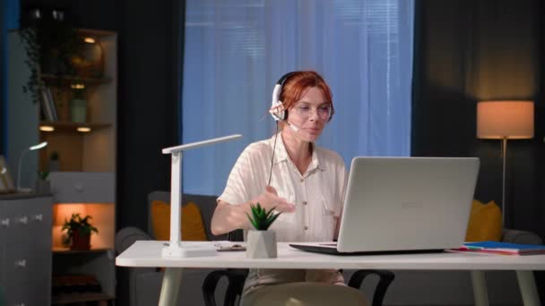 フリーランス 若い女性従業員はコールセンターでリモートで働き 自宅のコンピュータで座っている間 クライアントとヘッドセットを使用して話します — ストック動画