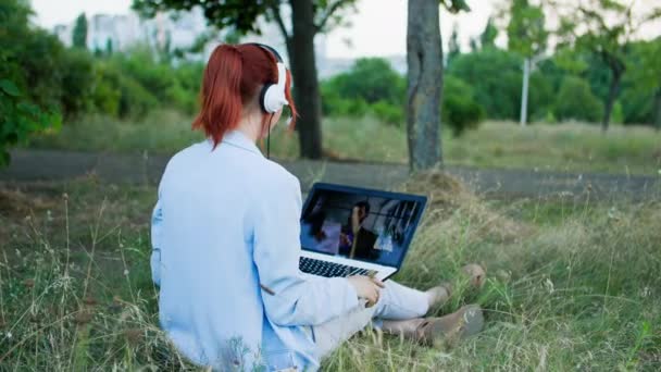 フリーランス 若い女性はヘッドセットを使用し 木の公園の背景に座っている間ラップトップ上のビデオリンクを介して通信します — ストック動画