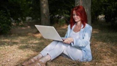 Serbest çalışan, genç bir iş kadını uzaktan çalışıyor ve parktaki bir klavyede ağaç kenarında oturuyor.