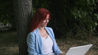 Evden işe giden, dışarıda dizüstü bilgisayarda çalışan parkta ağaçların yanında oturan genç bir kadın.