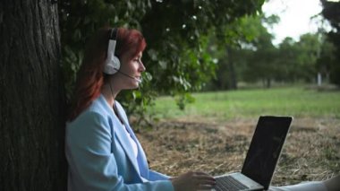 Serbest çalışan, kadın çalışan uzaktan çalışıyor ve ağaçların arasında park halinde otururken bir bilgisayara kulaklık ve telefon kullanarak müşteriye tavsiyelerde bulunuyor.