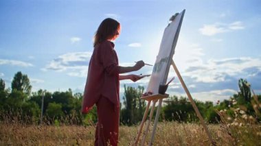 Fırça ve akrilik boya kullanan yetenekli bir kadın sanatçı açık hava sehpası kullanarak tuvale resim çiziyor.