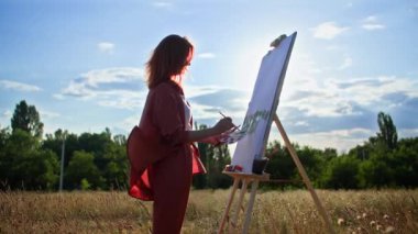 Fırça ve akrilik boya kullanan genç yaratıcı kadın ressam, gün ışığının arka planında bir sehpa kullanarak tuval üzerine resim çiziyor.