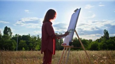 Hobi, yetenekli bir kadın, gün batımının sıcak yaz arkaplanında açık havada dururken resim çizer ve fırçayla resim yapar.
