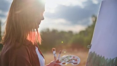 Yetenekli kadın ressam resim çizer. Şövaleyle resim çizer ve fırçayla resim yapar. Sıcak yaz gününün arka planında, güneş ışınları, siluet...