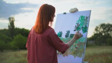 Elinde fırça olan genç bir kadın yeşil ağaçların arka planında sehpa kullanarak resim çiziyor.