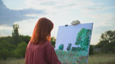 Yaratıcı bir hobi, genç bir kadın boya ve fırçalarla resim çizer, tuvale resim çizer ve gökyüzüne karşı park halinde durur.