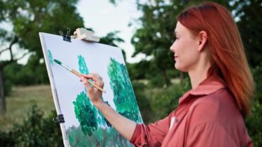 En sevdiği hobi, elinde boya ve fırça olan kadın sanatçı parktaki sehpayı kullanarak tuvale resim çizer, resme odaklanır.