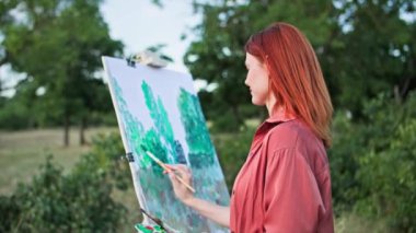 Resim ve fırça kullanan hoş bir kadın ağaçların arka planında dururken resim çiziyor.