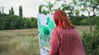 Açık hava hobisi, fırça ve resim kullanan yaratıcı genç bir kadın ağaçların arka planında bir resim çiziyor.